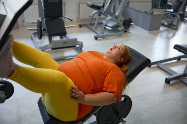 Spor salonunda bacak antrenmanı günü. Kilo vermeye çalışan tombul genç bir kadın sağlık kulübündeki spor malzemelerini kullanarak kalori yakmaya çalışıyor.