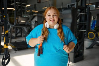 Spor kıyafetleri içinde mutlu ve şişman bir kadının portresi ve spor salonunda antrenman yaparken kameraya bakarak ip atlamak. Sportif sağlıklı yaşam tarzı ve obezitesiz yaşam seçimi.