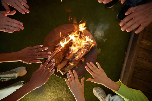 Group of young people warming hands over burning campfire. Closeup view on hands over outdoor fire