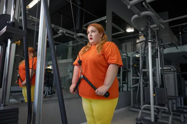 Exhausted excess overweight woman training with equipment at gym. Lovely pretty obese girl doing physical exercise for arms, shoulders and back. Sorts everyday routine for weightloss