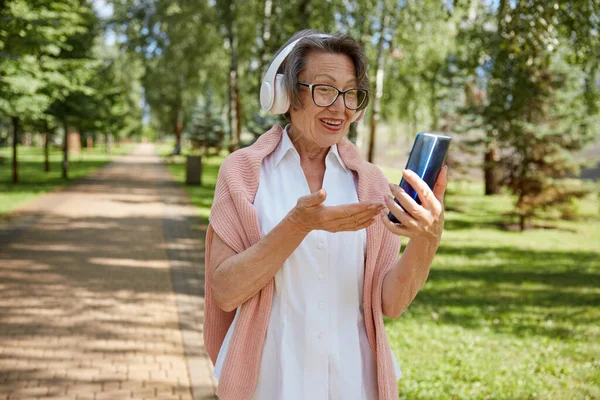 上了年纪的老妇人戴着耳机 一边在公园里散步 一边用无线网络与亲戚或朋友联系 在智能手机上进行在线视频通话 — 图库照片