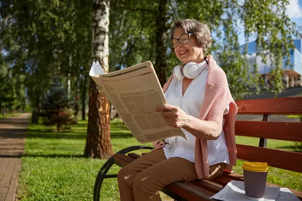 快乐地微笑着的老妇人坐在木制长椅上 在公园里看报纸 — 图库照片