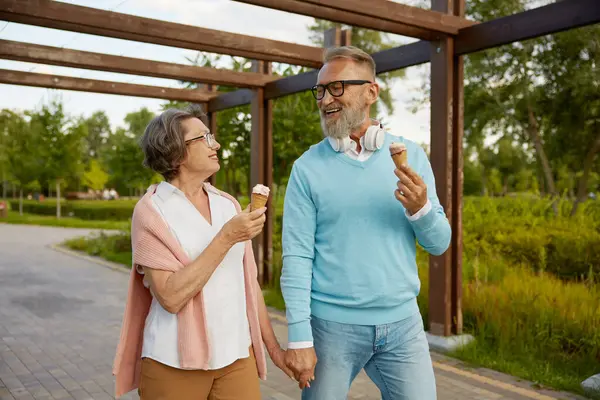 Cheerful senior couple eating ice-cream cone in park enjoying relax time. Joyful elderly lifestyle, pension time together concept