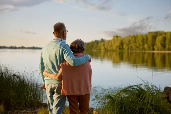Romantic calm senior couple looking faraway standing at river bank. Elderly people enjoying rest on nature feeling wellness and mindfulness
