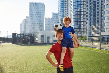 Futbol maçından sonra mutlu bir çocuğu omuzlarında tutan gülümseyen bir babanın portresi. Hafta sonları aile futbolu eğitimi, sağlıklı yaşam tarzı.