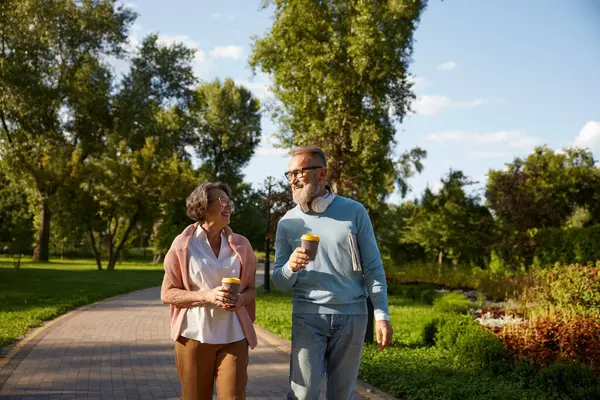 Happy elderly family having coffee break walking in park. Romantic aged mature man and woman looking happy enjoying healthy lifestyle on pension