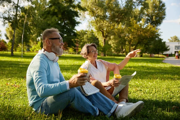 Senior loving couple drinking coffee and laughing while relaxing on green lawn grass outdoors in park