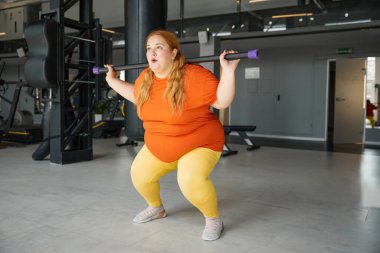 Kilolu bir kadın spor yapıyor. Vücut barı eğitim ekipmanlarıyla çömeliyor. Şişko kız güçlü, zayıf ve sağlıklı olmak için egzersiz yapıyor.