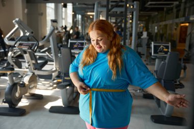 Tatmin olmuş kilolu bir kadın spor salonundaki zayıflama antrenmanından sonra ilk iyi sonucu ölçüyor.
