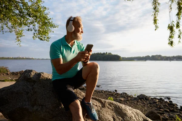 朝のトレーニングの後 川で休憩するシニアマン モチベーションのためにスマートフォンでお気に入りの音楽トラックを聴くヘッドセットを着用するアクティブな高齢者スポーツマン ロイヤリティフリーのストック写真