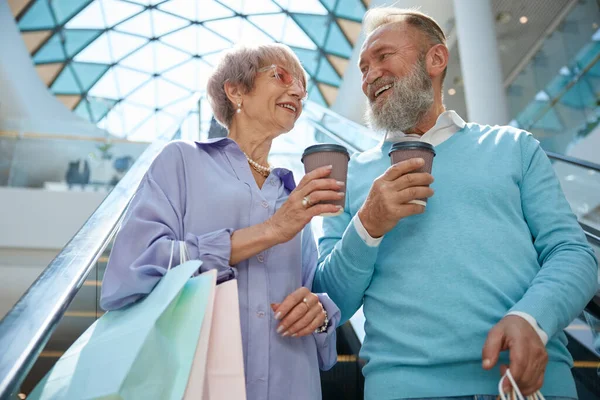 在商场自动扶梯上用购物袋和咖啡帽购物的老夫妇享受在一起的时光 — 图库照片