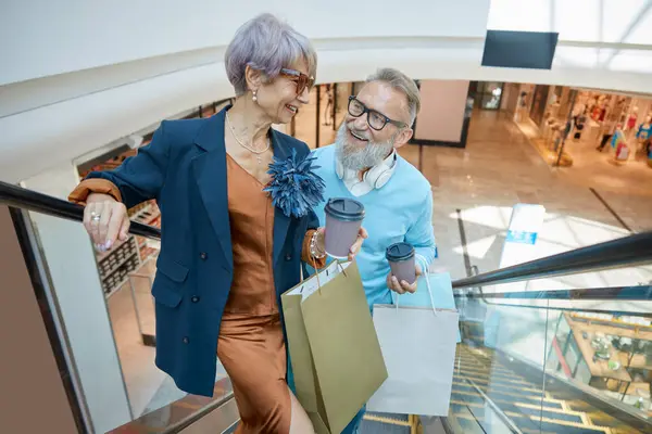Happy elderly couple buyers purchasing in mall for holidays. Senior wife and husband riding escalator sharing expression about shopping in new city market