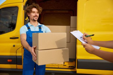 Dost canlısı gülümseyen genç kurye kontrol listesi ve imza için kalemle pano veriyor. Karton paketlerle dolu minibüsün yanında duran teslimatçı. Lojistik işçisi portresi