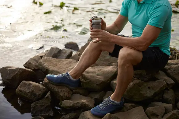Crop view of old mature sportsman in wild nature after morning jog. Senior man sitting on stone by pond with water lily flower drinking water and taking rest