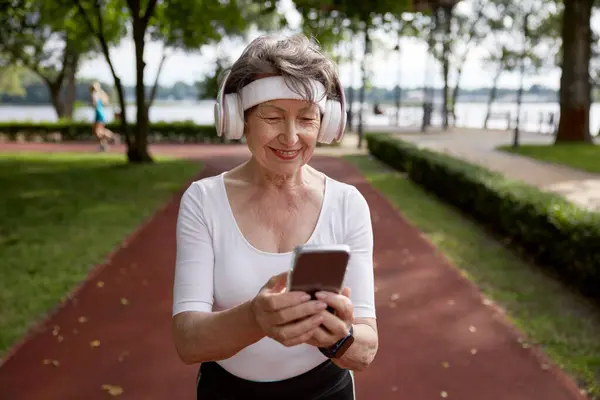 Elderly woman using mobile phone during running exercise outdoors at city park. Senior female browsing internet chatting with friends sharing good results of training