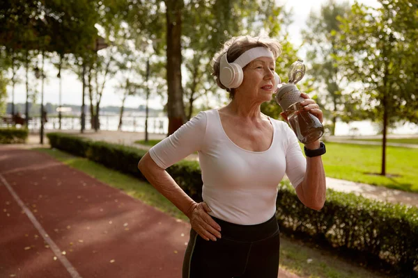Elderly woman drinking water taking short break during running exercise on stadium field at natural park. Sport hobby activity on retirement