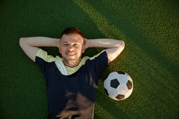 Happy smiling restful football player lying on green field grass overhead view. Sportsman feeling good and excited while rest on pitch