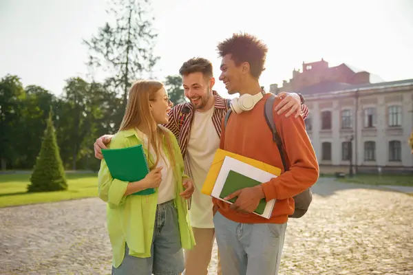 Multinational group of cheerful students hugging while chatting and laughing together at university campus. Interracial friendship, companionship, amity concept