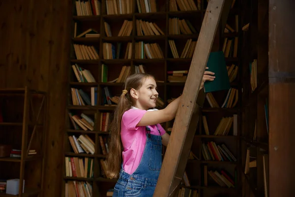 スマートガールの小学生が棚から本を取ります 教育のための教科書がたくさんある図書館のインテリアデザイン — ストック写真