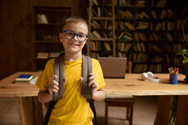 机のテーブルと本棚の背景を持つ家の図書館の上にバックパックを持つ小さな少年学生の肖像画 Eラーニングタイム — ストック写真