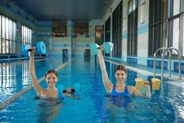 Young female friends doing aerobic workout in swimming pool. Two sportive women using dumbbells weights looking at camera