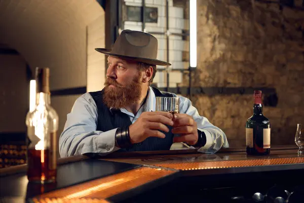 Portrait of mature gentleman in hat at bar counter. Elegant old-fashioned man drinking alcoholic beverage in bar