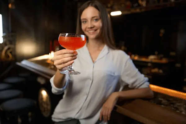 Relaxed cheerful young woman holding cocktail while sitting at bar counter looking at camera with smile