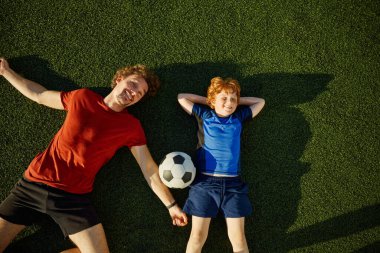 Baba ve küçük oğlu, futbol sahasındaki çimlerin üzerinde yatarken gülümsüyor ve eğleniyorlar. Mutlu aile açık hava eğlencesi.