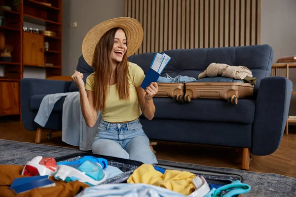 Happy young woman wearing straw beach hat and holding with flight tickets and passports dreaming in anticipation of upcoming trip on holiday vacation. Attractive packing suitcase for travel at home