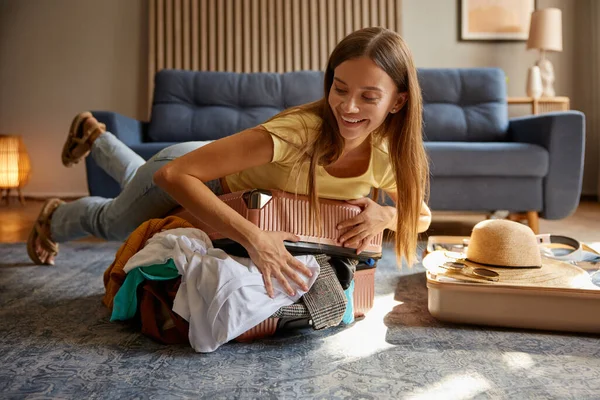 兴奋的女人在家里准备旅行箱 试图关上装满衣服的旅行箱 暑假准备概念 — 图库照片