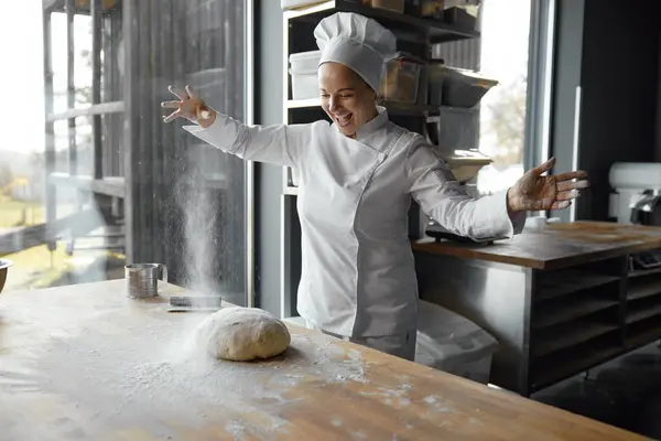 Excited enthusiastic young woman baker throwing flour on kneaded dough. Art of baking concept