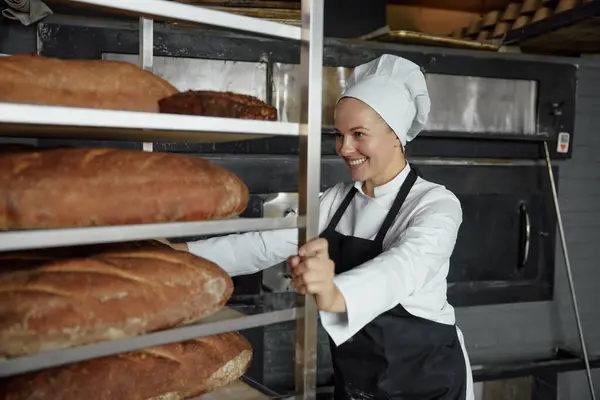 Experienced woman baker smiling happily pushing metal rack with fresh baked bread baguettes working in bakehouse