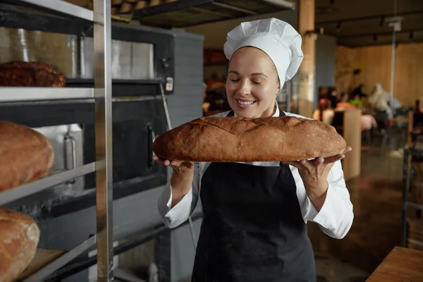 Smiling female baker smells freshly baked bread. Bakery shop worker