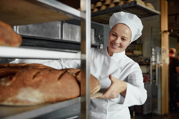 Woman baker smiling happily pushing wooden rack with fresh baked bread baguettes working in bakehouse