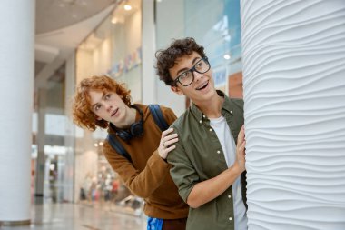 İki komik meraklı çocuk alışveriş merkezinin arkasındaki köşeden dışarı bakıyor. Neşeli öğrenci en iyi arkadaşlar süpermarkette eğleniyorlar.