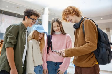 Farklı yaşlardaki mutlu çocuklar alışveriş merkezinde eğleniyor. Genç ve küçük çocuklar daire çizip telefon ekranına bakıyor. Bilgi arıyor, geziniyor ya da çekim üstüne fotoğraf izliyorlar.