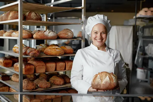 Happy woman baker chef holding bread standing near showcases with baked loaf. Bakery shop concept