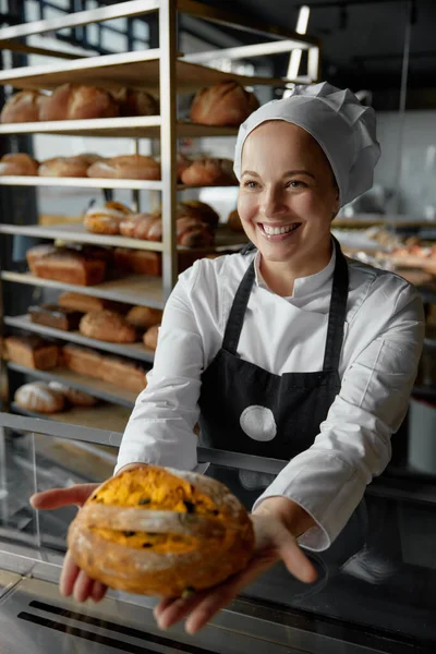 Happy woman baker chef holding bread standing near showcases with baked loaf. Bakery shop concept