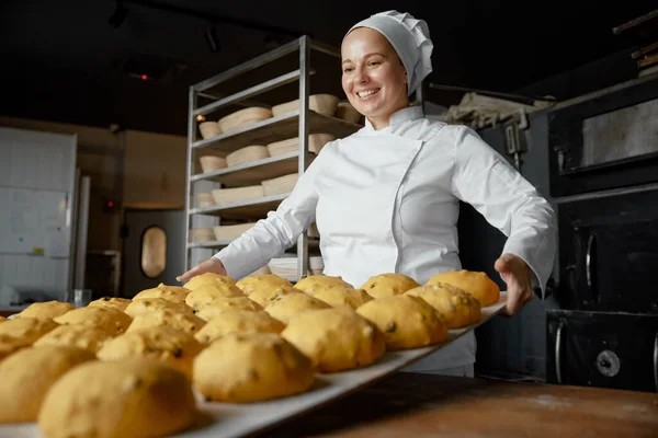 Satisfied positive woman baker at craft bakery workshop. Female chef in uniform looking on raw sweet pastry buns preparations on table in flour