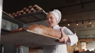 Kadın fırıncı, fırında çalışan taze ekmek ekmeğiyle ahşap askıları iterek mutlu bir şekilde gülümsüyor.