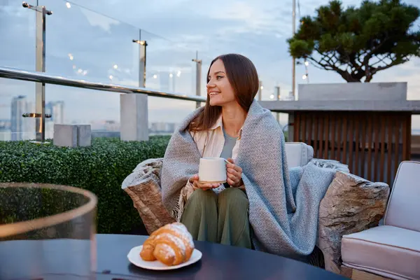 Young relaxed woman drinking coffee spending cozy evening at rooftop cafe. Millennial girl smiling enjoying panoramic view on cityscape sitting on home terrace. Rest and relax