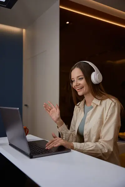 Young woman wearing headphones waving hand greeting hand gesture having meeting on laptop from home. Charming female freelancer holding business video conference or remote e-learning training class