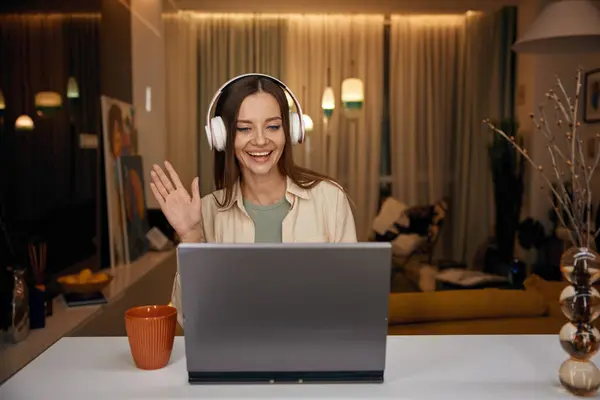 Woman wearing headphones waving hand greeting hand gesture having meeting on laptop from home. Charming female freelancer holding business video conference or remote e-learning training class