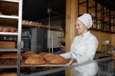 Fırıncı kadın, fırında çalışan taze ekmek ekmeğiyle metal rafları iterek mutlu bir şekilde gülümsüyor.