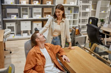 Modern mobilya galerisinde bilgisayar masasını seçen genç bir aile. Çekici bir eş kocaya sürpriz bir hediye hazırlıyor. Ev işyeri için masa alıyor.