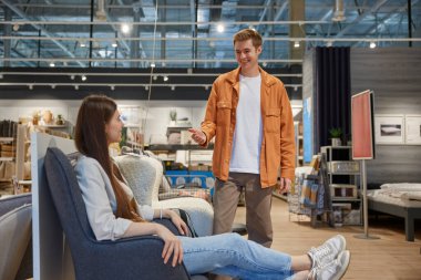 Genç bir çift, büyük indirimli ve çeşitli ürünler satan bir mağazada ev işleri için rahat bir sandalye seçiyor. Mobilya pazarında koltuğu test eden çekici bir kadın.