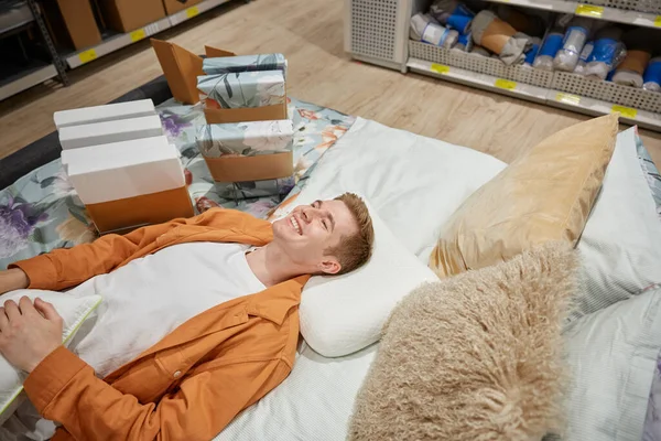 Mağaza mağazasında yastıkla yatakta uzanan yakışıklı, rahat bir adam. Genç adam alışveriş yaparken yatak odası için yeni yatak odası seçiyor.