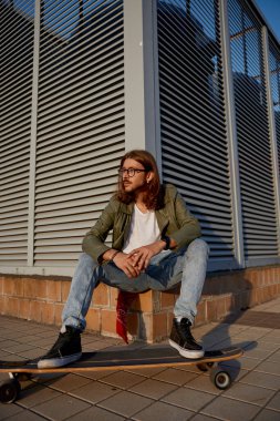 Şehir duvarında kaykayın üzerinde günlük giysiler giyen genç hippi. Moda tarzı, gençlik kültürü ve serbest stil