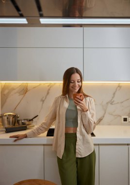 Genç, gülümseyen bir kadın kahve içiyor ve ev mutfağında sakin bir akşamın tadını çıkarıyor. Yüzünde tatmin edici bir ifade olan çekici bir kadın hayal kurarak geleceği planlamaya çalışıyor.