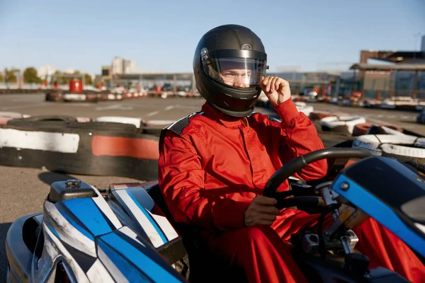 Handsome man driver wearing uniform taking off protective helmet while sitting at kart race car. Extreme go-kart training on race track
